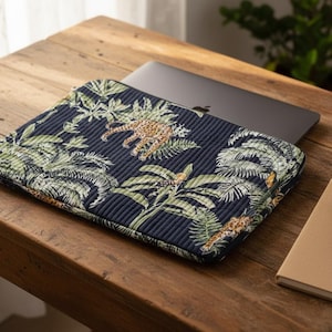 May include: A navy laptop sleeve with a jungle print featuring leopards and tropical foliage. The sleeve is on a wooden table with a laptop, notebook, and pen. The sleeve has a quilted texture.