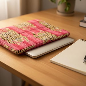 May include: A laptop computer is inside a pink and gold patterned laptop sleeve on a wooden desk. A spiral notebook and a black pen are next to the laptop. A plant and books are in the background.