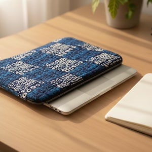 May include: A blue and white patterned laptop sleeve sits on a silver laptop. A notebook with a pen and a stack of books are also on the wooden desk. The sleeve has a quilted design.