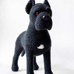 May include: A black crochet dog figurine with upright ears, a rounded snout, and a small white patch on its chest. The toy features dark brown plastic eyes and a black plastic nose, crafted with a textured yarn stitch pattern in a standing pose.