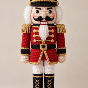 Könnte beinhalten: Eine gehäkelte Nussknacker-Puppe mit einem schwarzen Hut mit rotem Emblem, einer roten Jacke mit goldenen Verzierungen und Epauletten, einem schwarzen Gürtel mit goldener Schnalle, weißer Hose und schwarzen Stiefeln mit goldener Schnürung und weißem Bart.