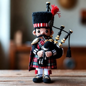 May include: A crocheted doll dressed as a Scottish bagpiper, wearing a black fuzzy busby hat with a red tassel, a red and green tartan kilt, and a black sporran. The figure holds a set of bagpipes with black and cream-colored pipes.