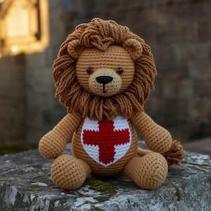 May include: A crocheted lion soft toy made from light brown yarn, featuring a thick textured mane and dark brown eyes. The lion has a white shield-shaped patch on its chest with a red cross stitched in the centre, and a small tufted tail.