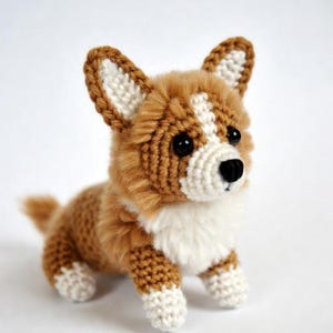 May include: A small, handcrafted crochet corgi dog figurine featuring a brown and white yarn body, pointed ears with white inner details, a white chest, and white paws. The plush toy has black plastic eyes and a black stitched nose.