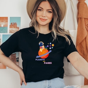 May include: A woman wearing a black t-shirt with a graphic of an astronaut sitting on a planet with the text "Planets and Pages".