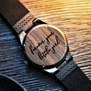 May include: A wooden wristwatch with a black leather band. The watch face is engraved with the words "forever your little girl".