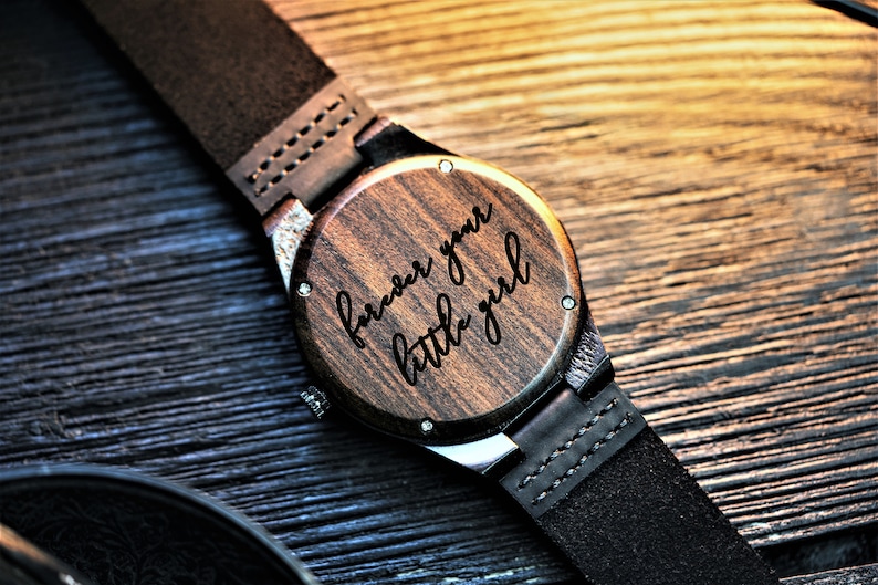 May include: A wooden wristwatch with a black leather band. The watch face is engraved with the words "forever your little girl".