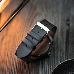 May include: A black leather watch band with a silver buckle. The band is attached to a wooden watch case.