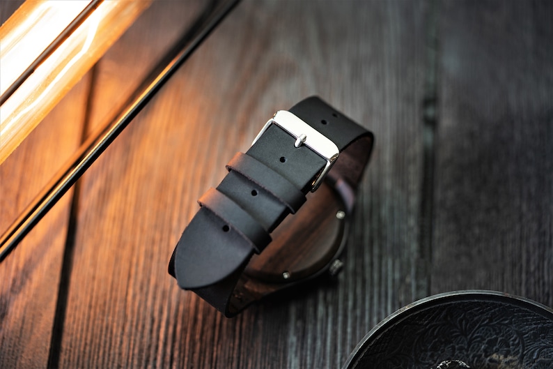 May include: A black leather watch band with a silver buckle. The band is attached to a wooden watch case.