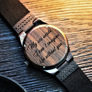 May include: A wooden wristwatch with a black leather band. The watch face is engraved with the words "My six words I can't imagine life without you."