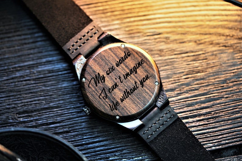 May include: A wooden wristwatch with a black leather band. The watch face is engraved with the words "My six words I can't imagine life without you."