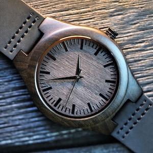 May include: A dark brown wooden wristwatch with a black leather band. The watch face has black hour and minute hands and black markings for the hours.