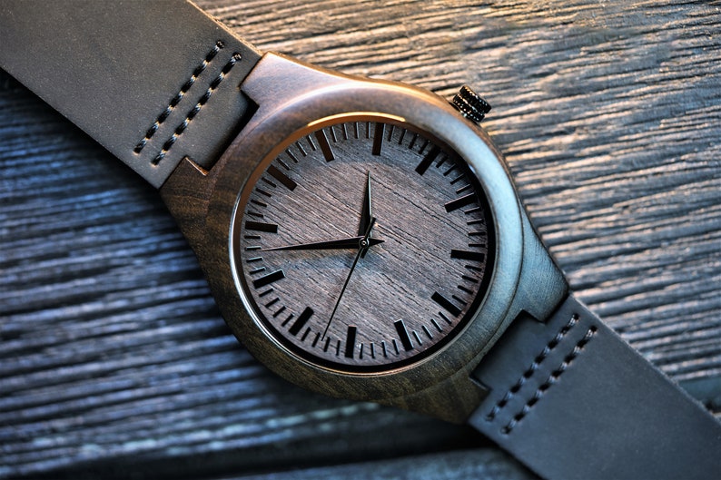 May include: A dark brown wooden wristwatch with a black leather band. The watch face has black hour and minute hands and black markings for the hours.