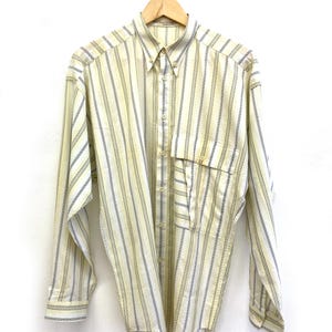 May include: A long-sleeve button-down shirt featuring a vertical striped pattern in pale yellow, white, and dark gray. The shirt includes a button-down collar, a single chest pocket with a flap, and buttoned cuffs, all displayed on a wooden hanger.