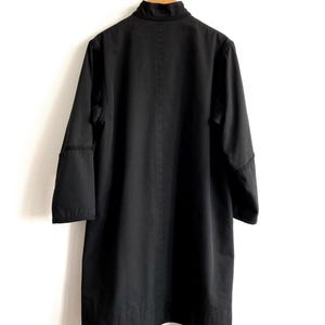 May include: A black coat hanging on a wooden hanger against a white wall. The coat has a high collar, long sleeves, and a straight, knee-length cut. The coat is made of a solid, dark fabric.