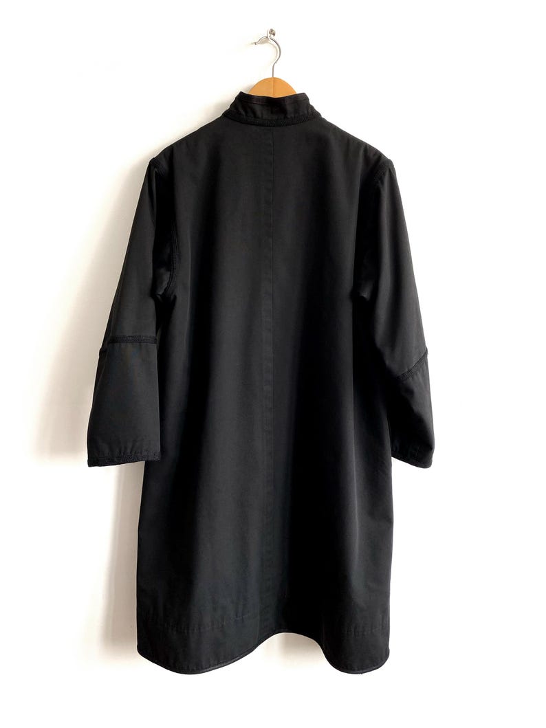 May include: A black coat hanging on a wooden hanger against a white wall. The coat has a high collar, long sleeves, and a straight, knee-length cut. The coat is made of a solid, dark fabric.