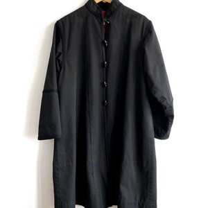 May include: A long, black coat with a mandarin collar and a row of buttons down the front. The coat has long sleeves and a red lining visible at the collar. It hangs on a wooden hanger against a white background.