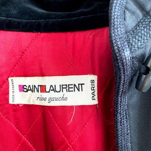 May include: Close-up of a vintage jacket interior. The jacket features a quilted red lining, a black velvet collar, and a label that reads "SAINT LAURENT rive gauche PARIS". The exterior is a dark gray fabric.
