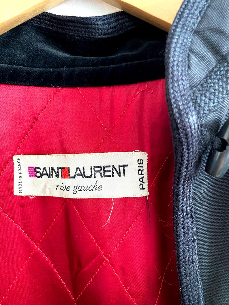 May include: Close-up of a vintage jacket interior. The jacket features a quilted red lining, a black velvet collar, and a label that reads "SAINT LAURENT rive gauche PARIS". The exterior is a dark gray fabric.