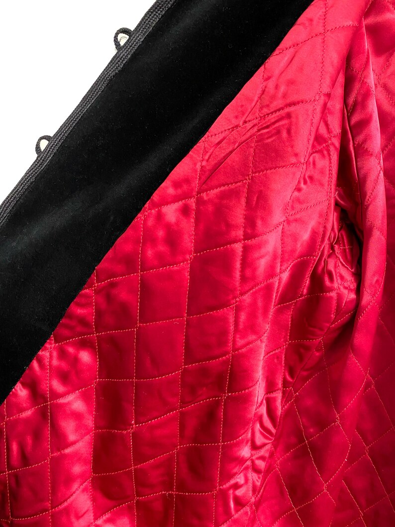 May include: Close-up of a black velvet jacket with a vibrant red quilted lining. The lining features a diamond pattern, and the jacket has black corded trim and small loops.