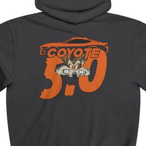 Könnte beinhalten: Dunkelgrauer Kapuzenpullover mit einem orangefarbenen Aufdruck. Der Aufdruck zeigt das Wort "COYOTE", eine Autosilhouette, die Zahl "5.0" und eine Cartoon-Kojotenfigur. Der Pullover hat eine Kapuze und lange Ärmel.