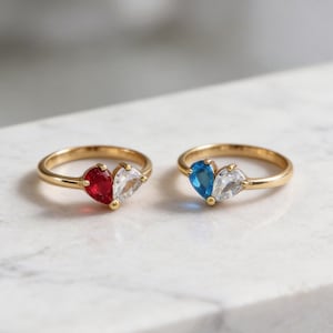 May include: Two gold rings, each with a heart-shaped design made of two gemstones. One ring has a red and clear stone combination, the other a blue and clear combination. The rings are displayed on a white marble surface.