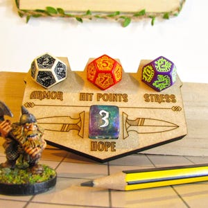 Może przedstawiać: Drewniany organizer do gier planszowych z kośćmi do gry dla Zbroi, Punktów Życia, Stresu i Nadziei. Zawiera miniaturową figurkę krasnoluda i ołówek. Kości są białe, czerwone i fioletowe. Organizer ma wygrawerowane słowa "Zbroja", "Punkty Życia", "Stres" i "Nadzieja".