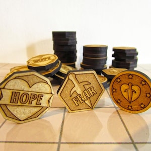 May include: Wooden tokens with laser-etched designs. Some tokens are round, others are octagonal. One token reads "HOPE" on a banner over a heart. Another reads "FEAR" with a sword. Stacks of black tokens are in the background.