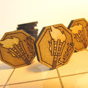 May include: Wooden tokens with a torch design and the word "Luck" etched on them. The tokens are hexagonal and appear to be made of a light brown wood. The torch has a flame at the top and a bundle of sticks below. Several tokens are visible.