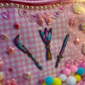 May include: Pink heart-shaped purse with a clear front, decorated with pink gingham fabric, bows, stars, and faux pearls. The purse features decorative charms and three weapon-shaped embellishments.