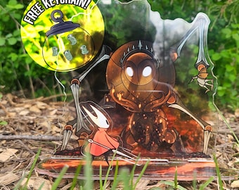 Puede incluir: Un expositor de acrílico transparente con un personaje vestido de rojo y máscara blanca frente a una gran criatura insectoide oscura con ojos brillantes. Una pegatina amarilla circular muestra el texto FREE KEYCHAIN! con un icono de frasco.