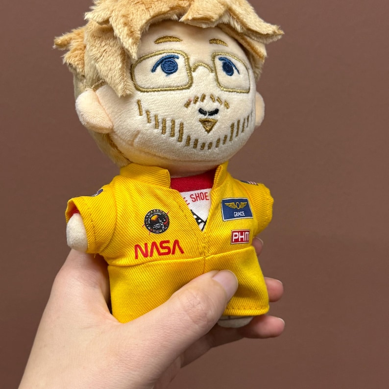 May include: A plush doll with blonde hair and glasses wearing a yellow NASA flight suit. The suit features embroidered patches including a NASA logo, a blue name tag reading GRACE, and a red PHM patch. The doll has a stitched beard and mustache.