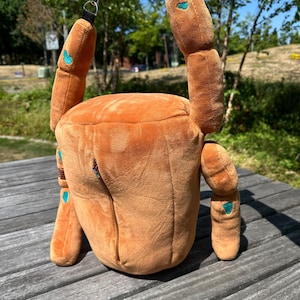 May include: A plush, orange, crab-shaped bag featuring a soft, textured exterior with small teal accents on the limbs. The bag includes a vertical zipper closure on the front and is equipped with black straps and metal hardware for carrying.