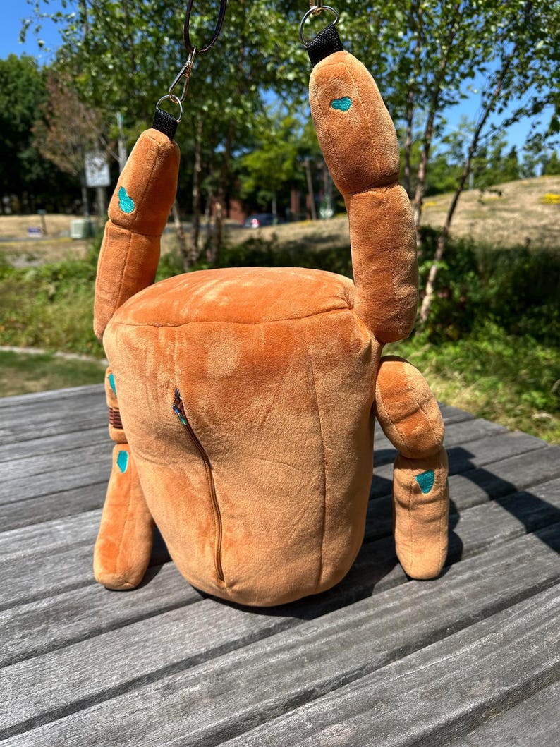 May include: A plush, orange, crab-shaped bag featuring a soft, textured exterior with small teal accents on the limbs. The bag includes a vertical zipper closure on the front and is equipped with black straps and metal hardware for carrying.