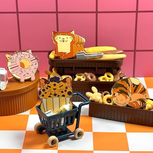 May include: A whimsical display of cat-themed enamel pins and miniature bakery items. The pins include a donut, a loaf of bread, a cupcake, and a sleeping cat. A miniature shopping cart is filled with tiny items. The background features a pink wall and an orange and white checkered floor.