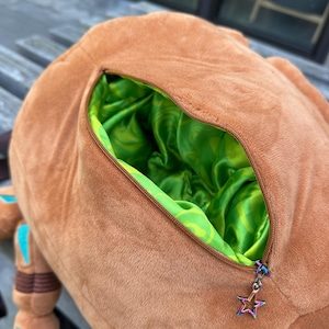 May include: A soft brown plush toy featuring a zippered opening that reveals a vibrant lime green satin lining with a swirling pattern. The zipper pull is a small, iridescent metal star charm attached to the brown fabric exterior.