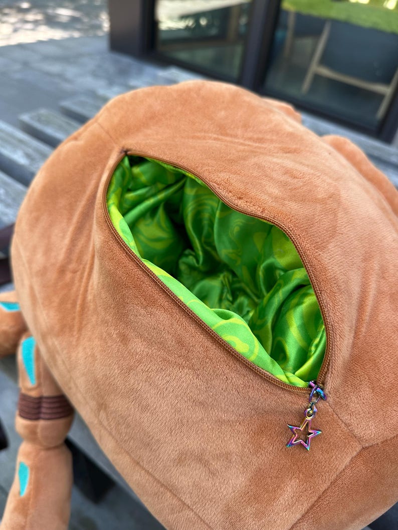 May include: A soft brown plush toy featuring a zippered opening that reveals a vibrant lime green satin lining with a swirling pattern. The zipper pull is a small, iridescent metal star charm attached to the brown fabric exterior.
