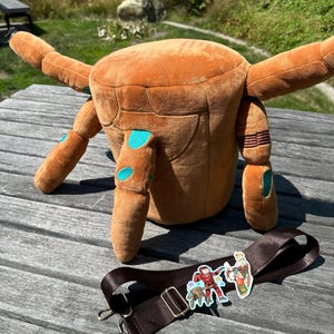 Puede incluir: Un bolso de peluche color canela con forma de criatura de cuatro extremidades extendidas, con detalles ovalados en color verde azulado en las patas. Incluye una correa ajustable de color marrón oscuro con dos pegatinas de personajes.