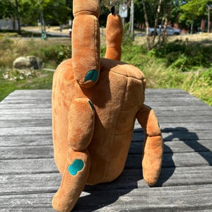 May include: A plush, tan-colored bag shaped like a crab, featuring soft fabric, two upright pincers, and four extended legs. The crab has small, teal-colored circular accents on its joints and a dark brown strap attached to the top for carrying.