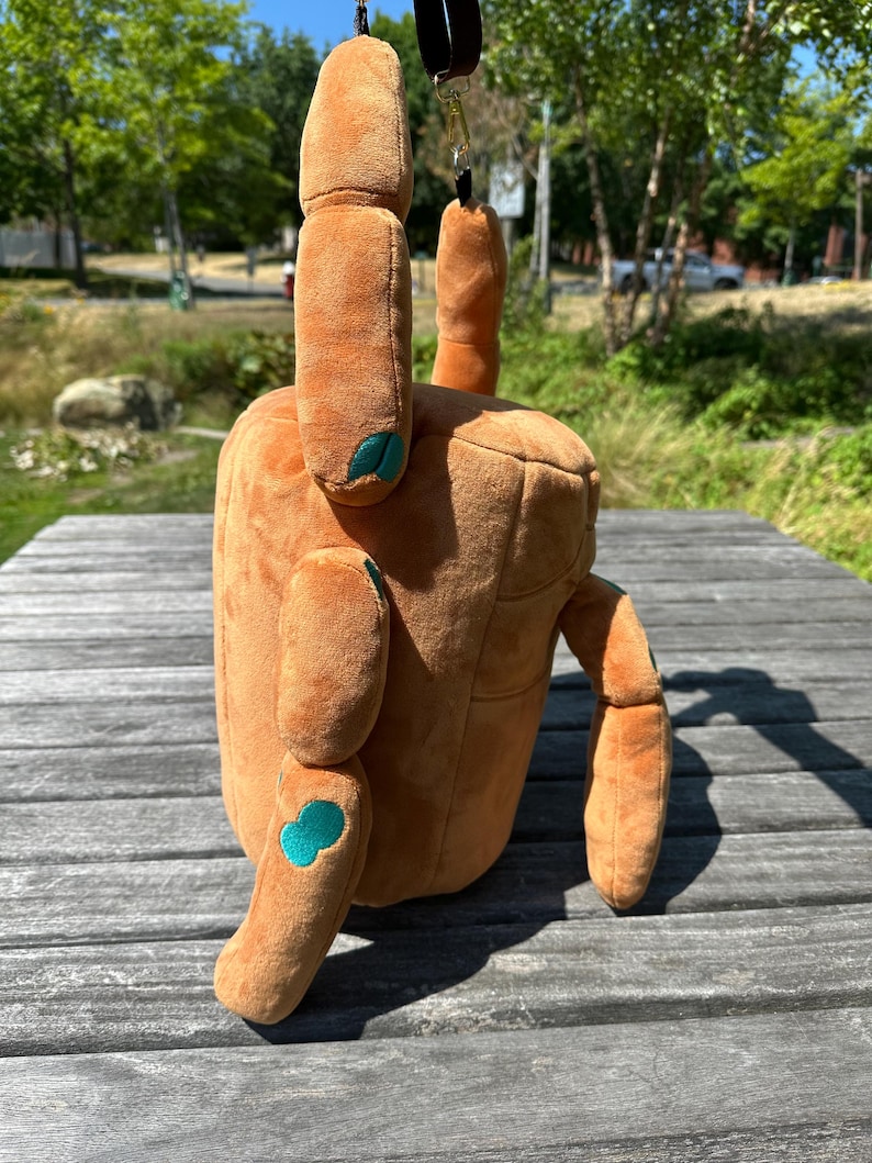 May include: A plush, tan-colored bag shaped like a crab, featuring soft fabric, two upright pincers, and four extended legs. The crab has small, teal-colored circular accents on its joints and a dark brown strap attached to the top for carrying.
