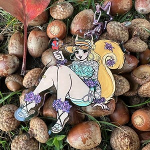 Op de afbeelding: Een kleurrijke acryl bedel met een stripfiguur met blond haar, een hoed en een turquoise top, versierd met paarse bloemen. De bedel is bevestigd aan een zilverkleurige sleutelhanger met een stervormige bedel. De achtergrond bevat eikels en gras.