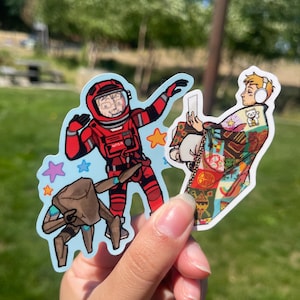 May include: Two die-cut vinyl stickers. One sticker features a person in a red NASA spacesuit with a brown robotic creature and colorful stars. The second sticker shows a person wearing white headphones wrapped in a colorful patterned quilt.