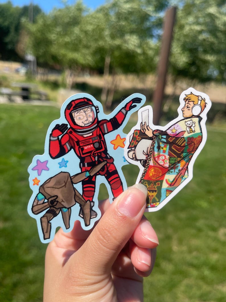 May include: Two die-cut vinyl stickers. One sticker features a person in a red NASA spacesuit with a brown robotic creature and colorful stars. The second sticker shows a person wearing white headphones wrapped in a colorful patterned quilt.
