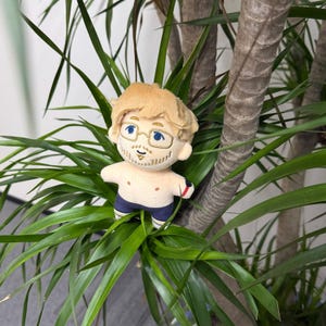 May include: A plush doll featuring a character with light brown hair, embroidered blue eyes, and gold-rimmed glasses. The figure has a light skin tone, a short beard, and is wearing dark blue shorts with white and yellow trim and a red arm band.