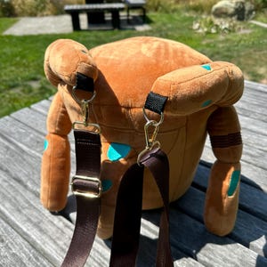 May include: A plush brown backpack featuring a rounded shape with teal spots and two dangling arms. The bag includes adjustable dark brown nylon straps attached with gold-toned metal lobster clasps and hardware, providing a functional design.