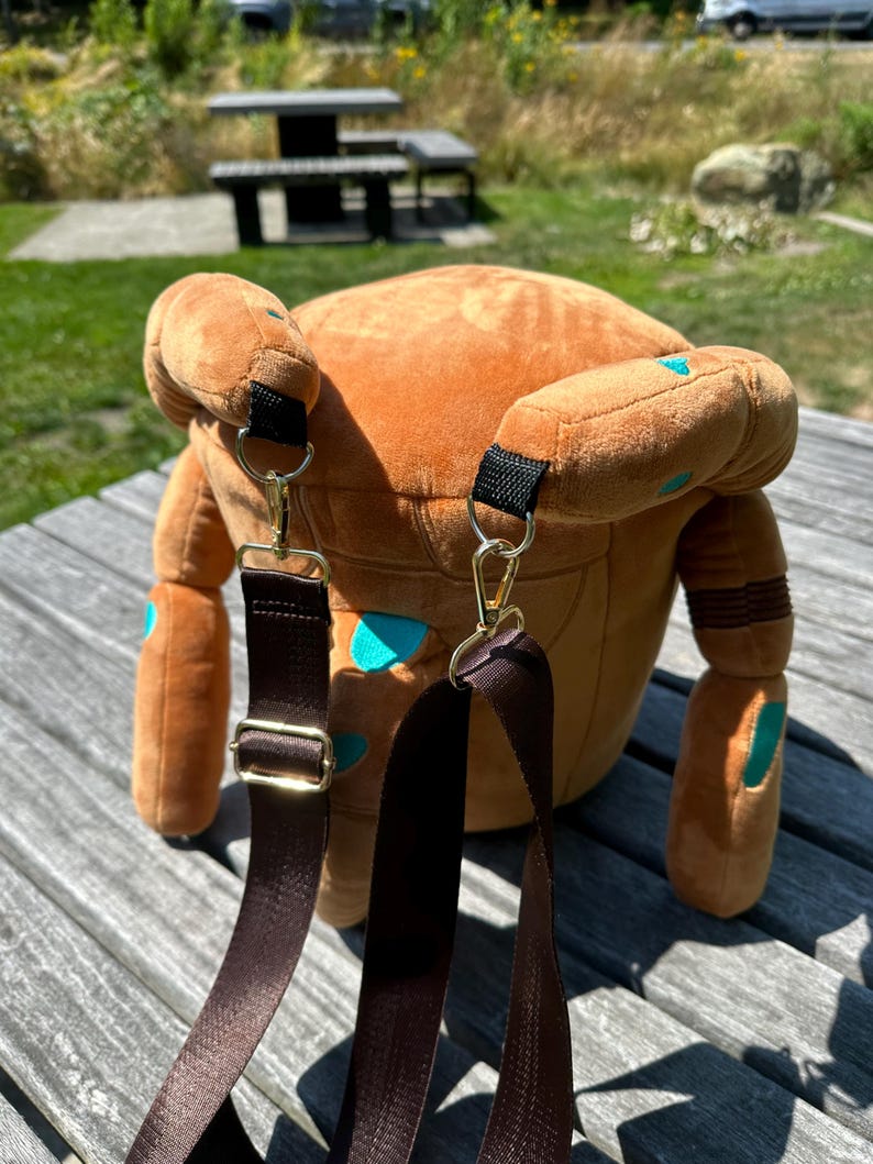May include: A plush brown backpack featuring a rounded shape with teal spots and two dangling arms. The bag includes adjustable dark brown nylon straps attached with gold-toned metal lobster clasps and hardware, providing a functional design.