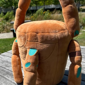 May include: A plush, brown, cylindrical bag shaped like a crab with articulated limbs. The soft fabric features teal-colored spots on the legs and body, with a black strap attached to the top. The design includes segmented limbs and a flat top.