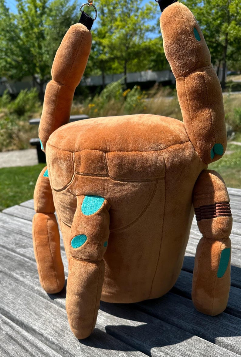 May include: A plush, brown, cylindrical bag shaped like a crab with articulated limbs. The soft fabric features teal-colored spots on the legs and body, with a black strap attached to the top. The design includes segmented limbs and a flat top.