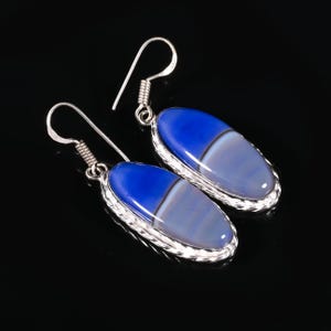 May include: A pair of oval earrings featuring blue and grey stones. The earrings have a silver-coloured metal border and hook. The blue is a deep, vibrant shade, and the grey has a striped pattern. The background is black.