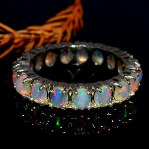 May include: A silver ring featuring a series of oval-cut opals. The opals display a play of color with flashes of blue, orange, and green. The ring is set against a dark background, highlighting the gemstones.